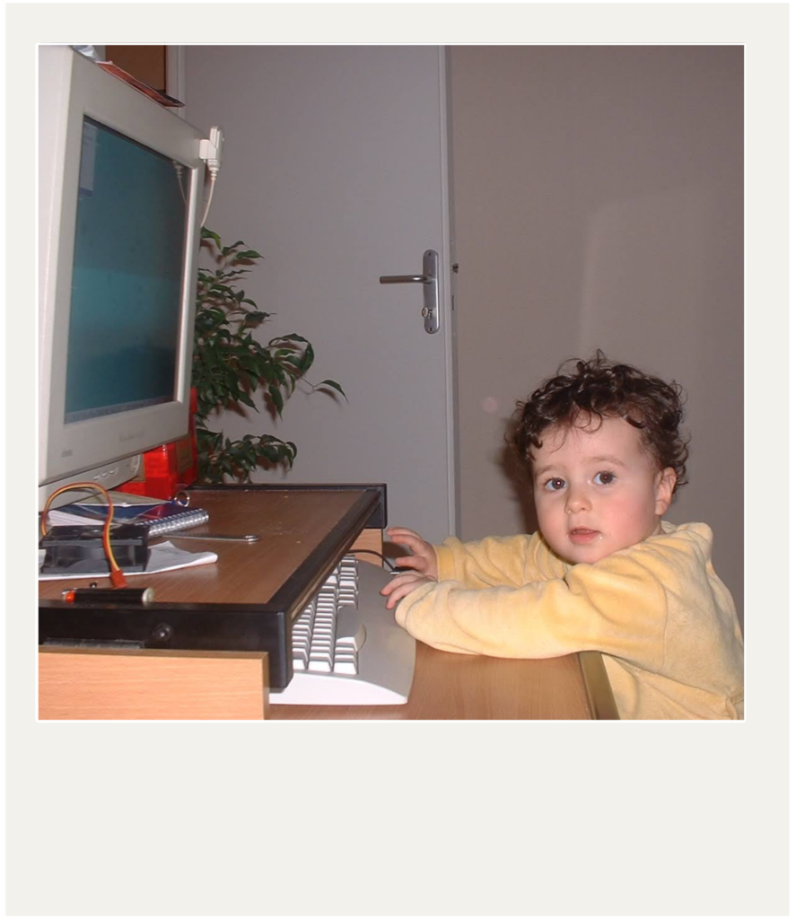A young child with curly hair and wearing a pale yellow bodysuit sits at a light-colored wooden desk, facing a '90s desktop computer. She places her hands on the white keyboard and looks up at the camera with a curious expression. The photo has a white Polaroid-like frame.