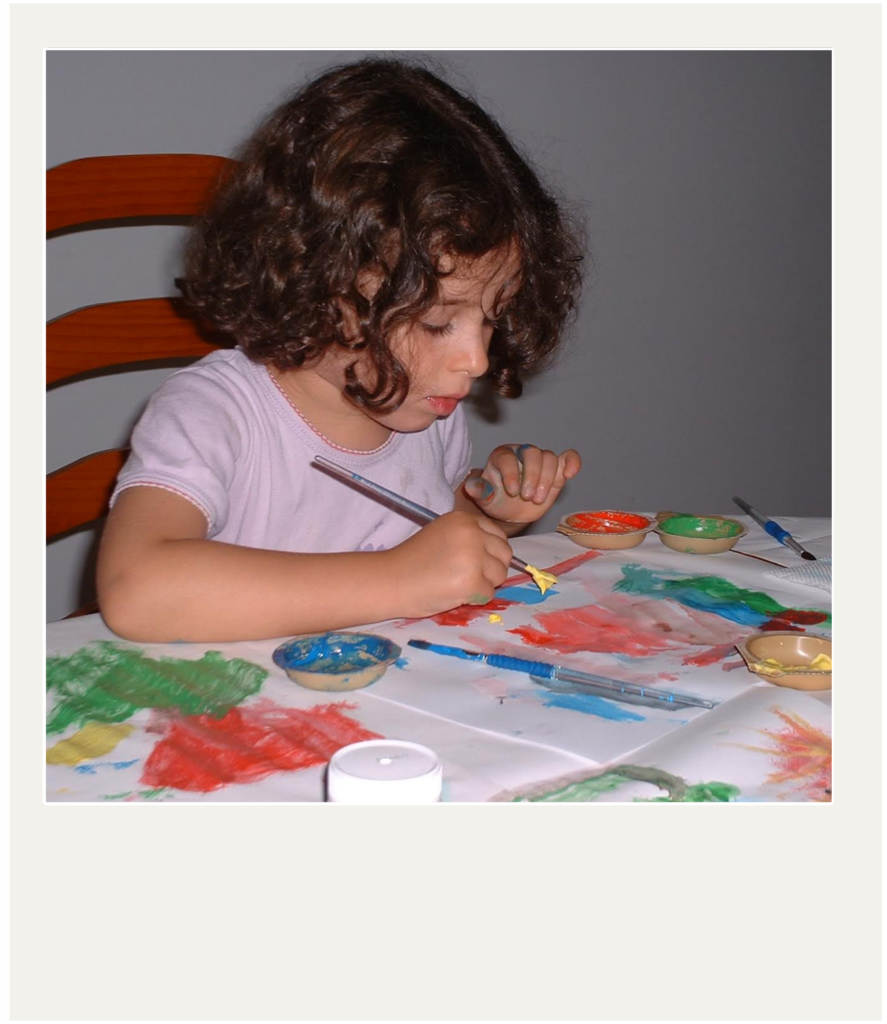 A young child sits at a table, painting. They are seated in a wooden chair and use a paintbrush to apply yellow paint to a piece of paper. Several paint containers and another brush are scattered around their artwork. Their fingers are smudged with paint. The image has a white, Polaroid-style border.
