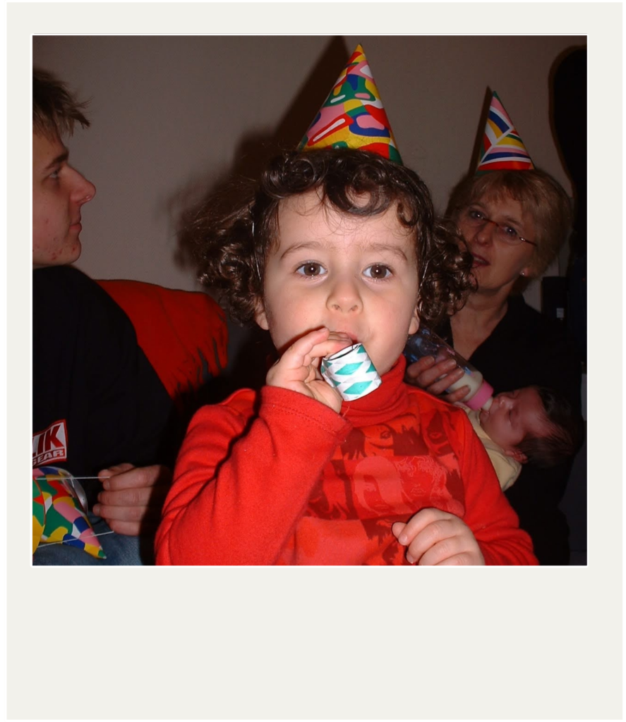 A young child with curly hair, wearing a colorful party hat, blows a green and white party horn.