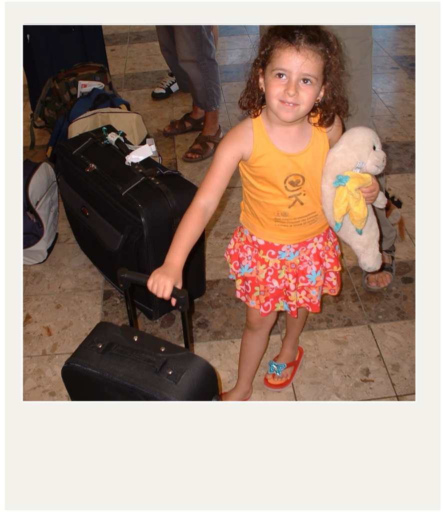 A young girl stands in what appears to be an airport, holding the handle of a small black wheeled suitcase. She's wearing an orange tank top, a red and orange floral skirt and orange flip-flops. She's holding a white stuffed seal with a yellow and blue scarf. Behind her, a larger black suitcase and other luggage can be seen. Parts of the legs and feet of several other people are also visible in the background. The floor is covered with beige and brown tiles. The image has a white frame, like a Polaroid photo.